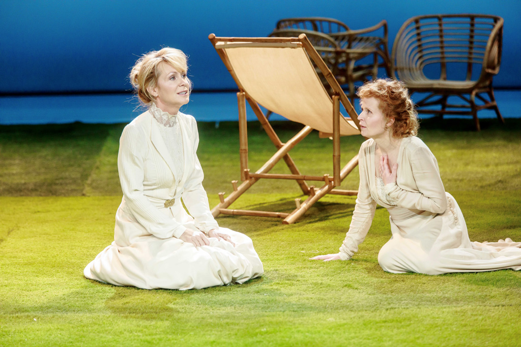Zdjęcie ze spektaklu. Dwie kobiety w białych sukniach siedzą na trawie, rozmawiają.